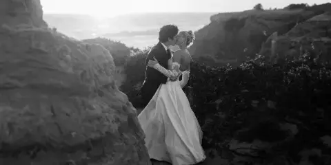 A black and white photograph of a bride and groom kissing on a rocky coastal cliff.