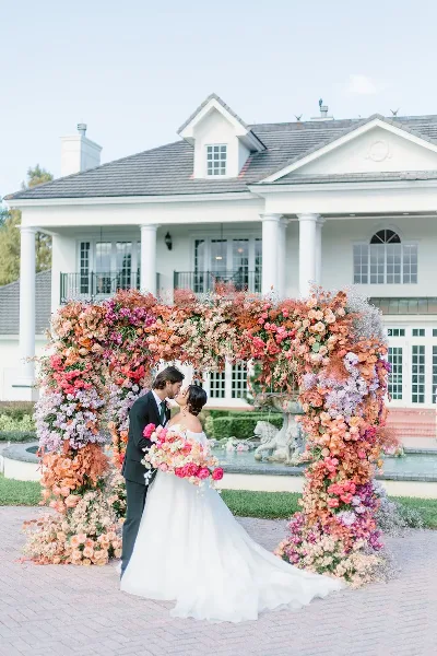 A bride and groom standing in front of a large, colorful floral arch installation at a wedding venue.