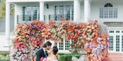 A bride and groom standing in front of a large, colorful floral arch installation at a wedding venue.