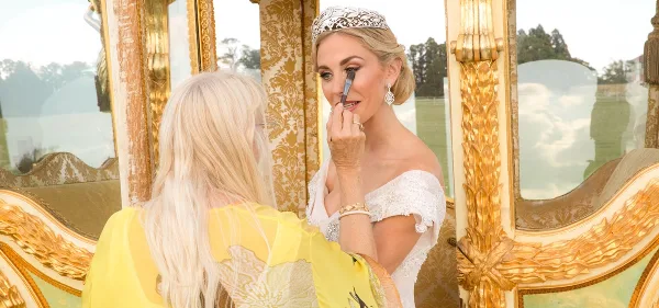 A makeup artist applying makeup to a bride wearing a tiara in front of a gold-framed mirror.