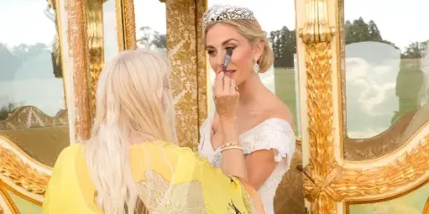 A makeup artist applying makeup to a bride wearing a tiara in front of a gold-framed mirror.