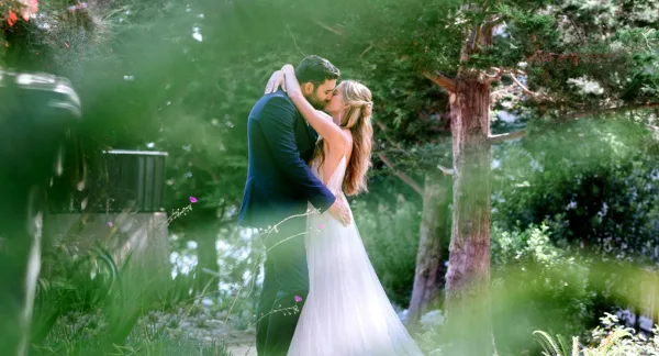 A bride and groom sharing a kiss in a lush, green outdoor garden setting.