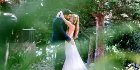 A bride and groom sharing a kiss in a lush, green outdoor garden setting.