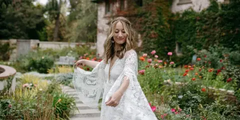 A bride with styled hair and makeup wearing a white lace dress in a garden setting.