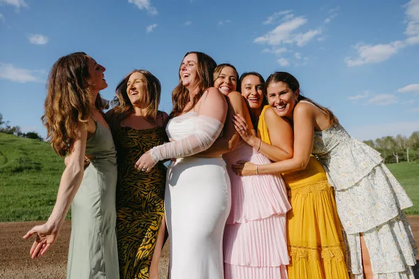 A bride in a white gown and five bridesmaids in colorful dresses laughing together outdoors on a sunny day.