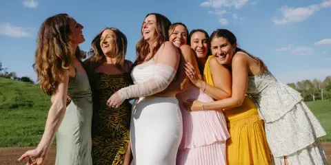 A bride in a white gown and five bridesmaids in colorful dresses laughing together outdoors on a sunny day.