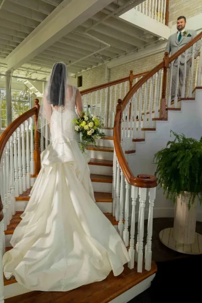 A bride in a white gown and veil walks down a wooden staircase toward a groom in a grey suit.