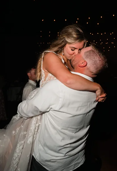 A groom lifts his bride for a kiss during a first dance at a wedding reception, illuminated by string lights in the background.