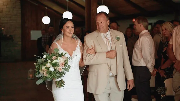 A bride in a white lace gown walks arm-in-arm with her father in a beige suit during a wedding ceremony, holding a bouquet of roses and greenery.
