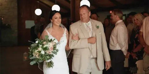 A bride in a white lace gown walks arm-in-arm with her father in a beige suit during a wedding ceremony, holding a bouquet of roses and greenery.