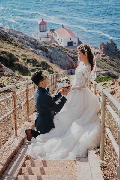 A groom kneeling on one knee in front of a bride in a white wedding dress, set against a coastal background with a lighthouse.