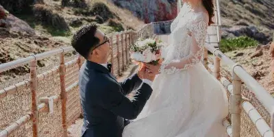 A groom kneeling on one knee in front of a bride in a white wedding dress, set against a coastal background with a lighthouse.