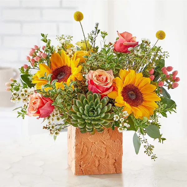 A vibrant floral arrangement featuring yellow sunflowers, peach roses, and greenery in a textured terracotta vase.