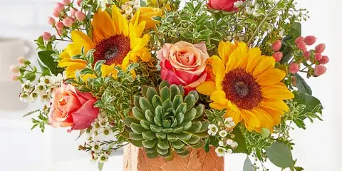 A vibrant floral arrangement featuring yellow sunflowers, peach roses, and greenery in a textured terracotta vase.