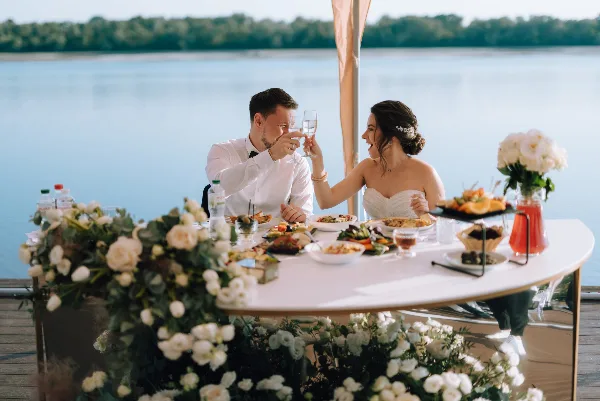 A bride and groom toasting with wine glasses at a wedding reception table set with food and flowers.