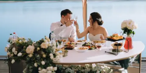 A bride and groom toasting with wine glasses at a wedding reception table set with food and flowers.