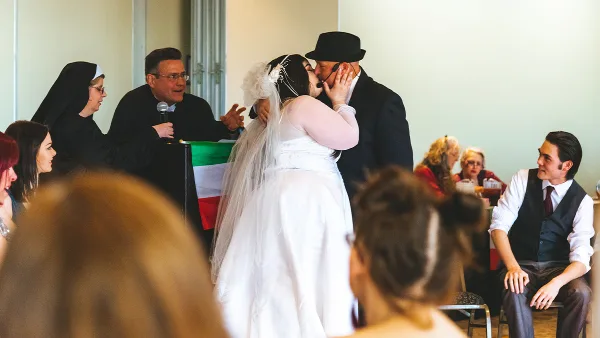 A bride and groom sharing a kiss during a wedding ceremony in a large indoor hall with guests seated in the background.