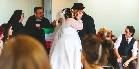 A bride and groom sharing a kiss during a wedding ceremony in a large indoor hall with guests seated in the background.