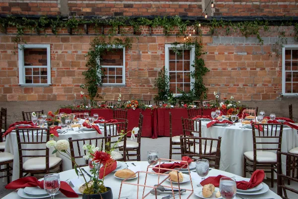 An outdoor wedding reception space featuring round tables with white linens, red napkins, and floral centerpieces set against a brick wall.