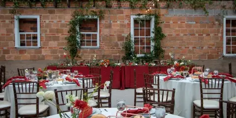 An outdoor wedding reception space featuring round tables with white linens, red napkins, and floral centerpieces set against a brick wall.