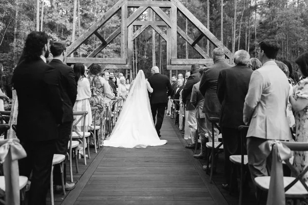 Black-and-white photo of a bride walking down a wooden aisle toward an open-air timber chapel surrounded by trees, with seated guests on either side.