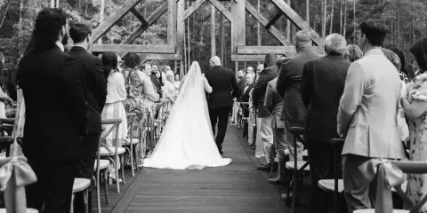 Black-and-white photo of a bride walking down a wooden aisle toward an open-air timber chapel surrounded by trees, with seated guests on either side.