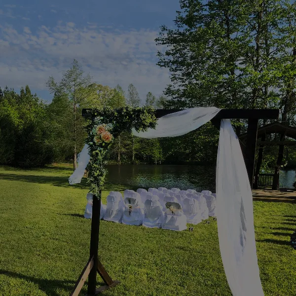 A wedding ceremony setup with a floral arch and white chairs on a grassy lawn next to a pond and wooden gazebo.