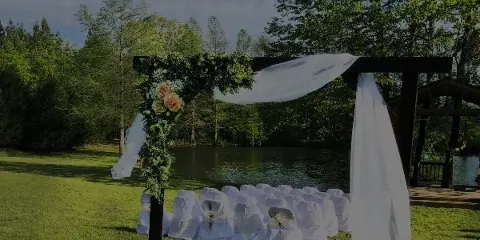 A wedding ceremony setup with a floral arch and white chairs on a grassy lawn next to a pond and wooden gazebo.
