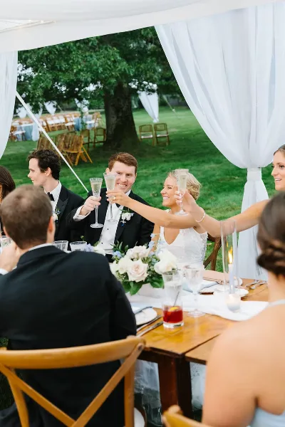 A bride and groom toasting with champagne flutes at a wedding reception table set with white flowers and candles under a white tent.