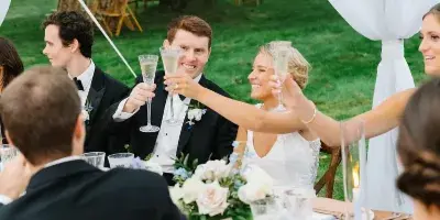 A bride and groom toasting with champagne flutes at a wedding reception table set with white flowers and candles under a white tent.