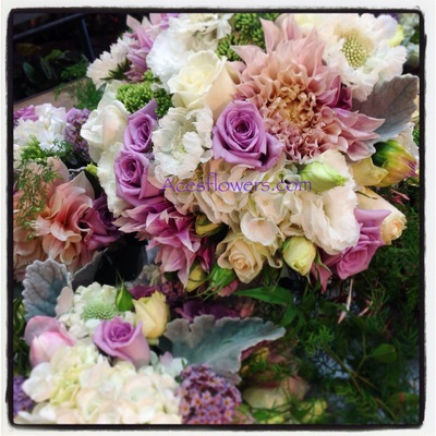 A close-up photograph of a lush floral arrangement with purple roses, white hydrangeas, and pink dahlias.