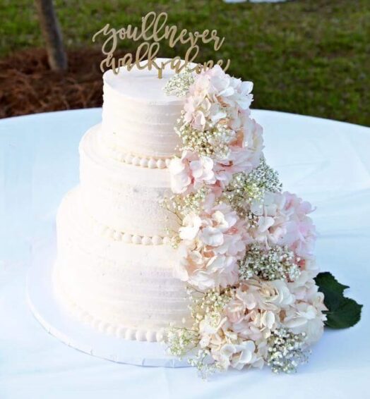 A three-tiered white wedding cake decorated with pink and white flowers and a gold topper.