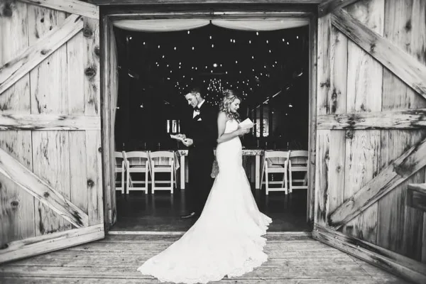 A black and white photo of a bride and groom standing in the doorway of a rustic wooden barn with string lights and chairs visible inside.