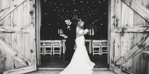 A black and white photo of a bride and groom standing in the doorway of a rustic wooden barn with string lights and chairs visible inside.