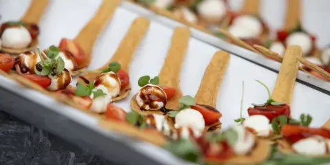 A close-up of a catering buffet featuring rows of appetizers served on wooden spoons, garnished with fresh herbs and balsamic glaze.