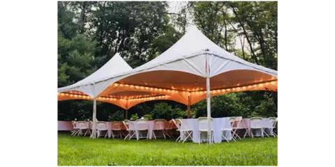 A white high-peak tent set up on a grassy lawn with string lights, tables, and chairs arranged for an event.