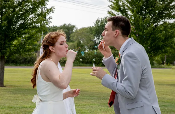 A bride and groom in wedding attire blowing bubbles together in a grassy field.