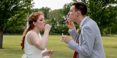 A bride and groom in wedding attire blowing bubbles together in a grassy field.