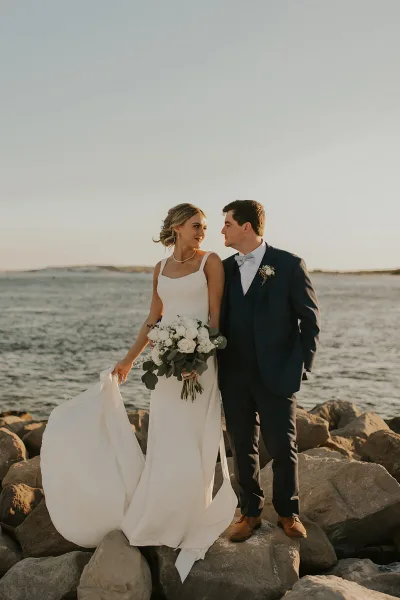 A bride and groom standing on rocks by the water, looking at each other.