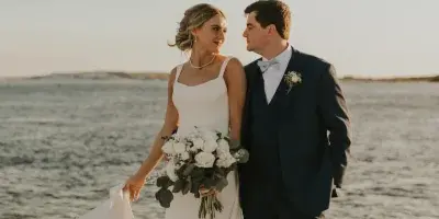 A bride and groom standing on rocks by the water, looking at each other.