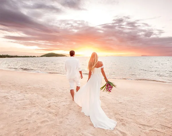 A bride and groom walking on a sandy beach towards a sunset over the ocean.