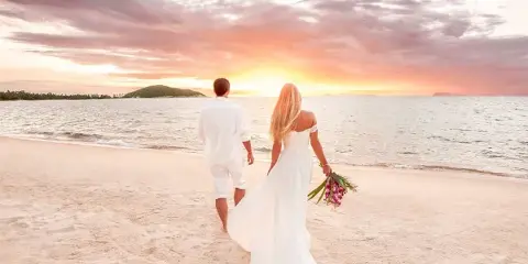 A bride and groom walking on a sandy beach towards a sunset over the ocean.