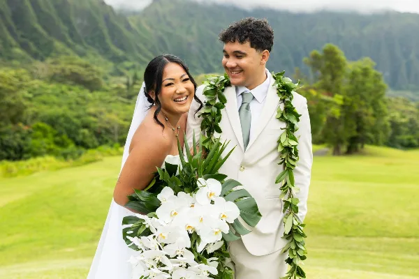 A smiling bride and groom wearing leis and holding a white orchid bouquet, standing outdoors with a green mountainous landscape in the background.