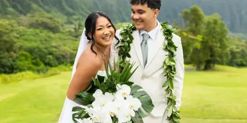 A smiling bride and groom wearing leis and holding a white orchid bouquet, standing outdoors with a green mountainous landscape in the background.