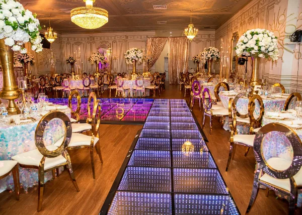A wedding venue interior featuring a long LED dance floor, gold chairs, and white floral centerpieces.