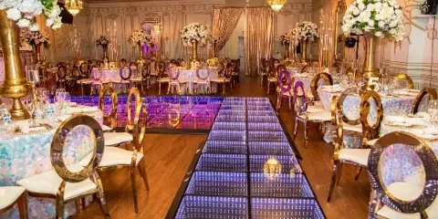 A wedding venue interior featuring a long LED dance floor, gold chairs, and white floral centerpieces.