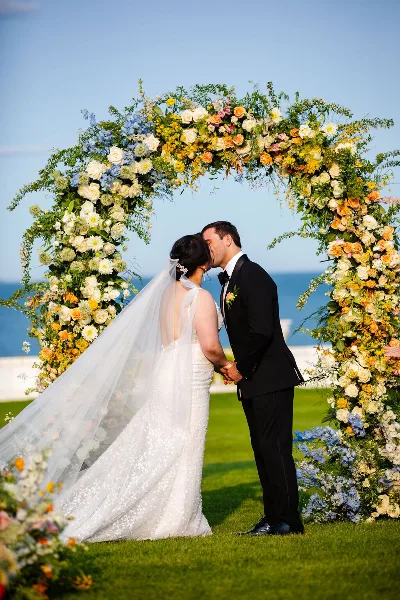 A bride and groom kissing under a large, colorful floral arch installation with a coastal background.