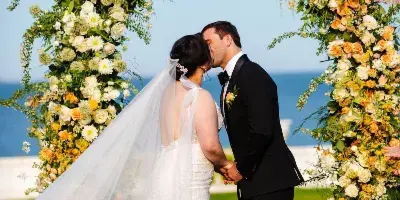 A bride and groom kissing under a large, colorful floral arch installation with a coastal background.