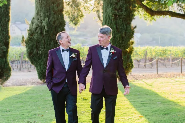 Two men in maroon tuxedos walking hand-in-hand through a vineyard during a wedding.