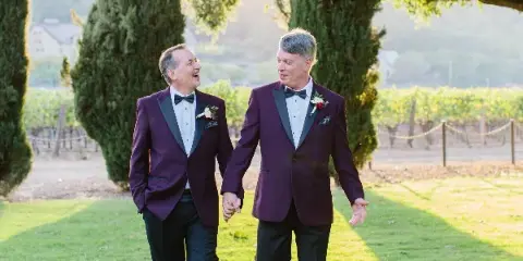 Two men in maroon tuxedos walking hand-in-hand through a vineyard during a wedding.